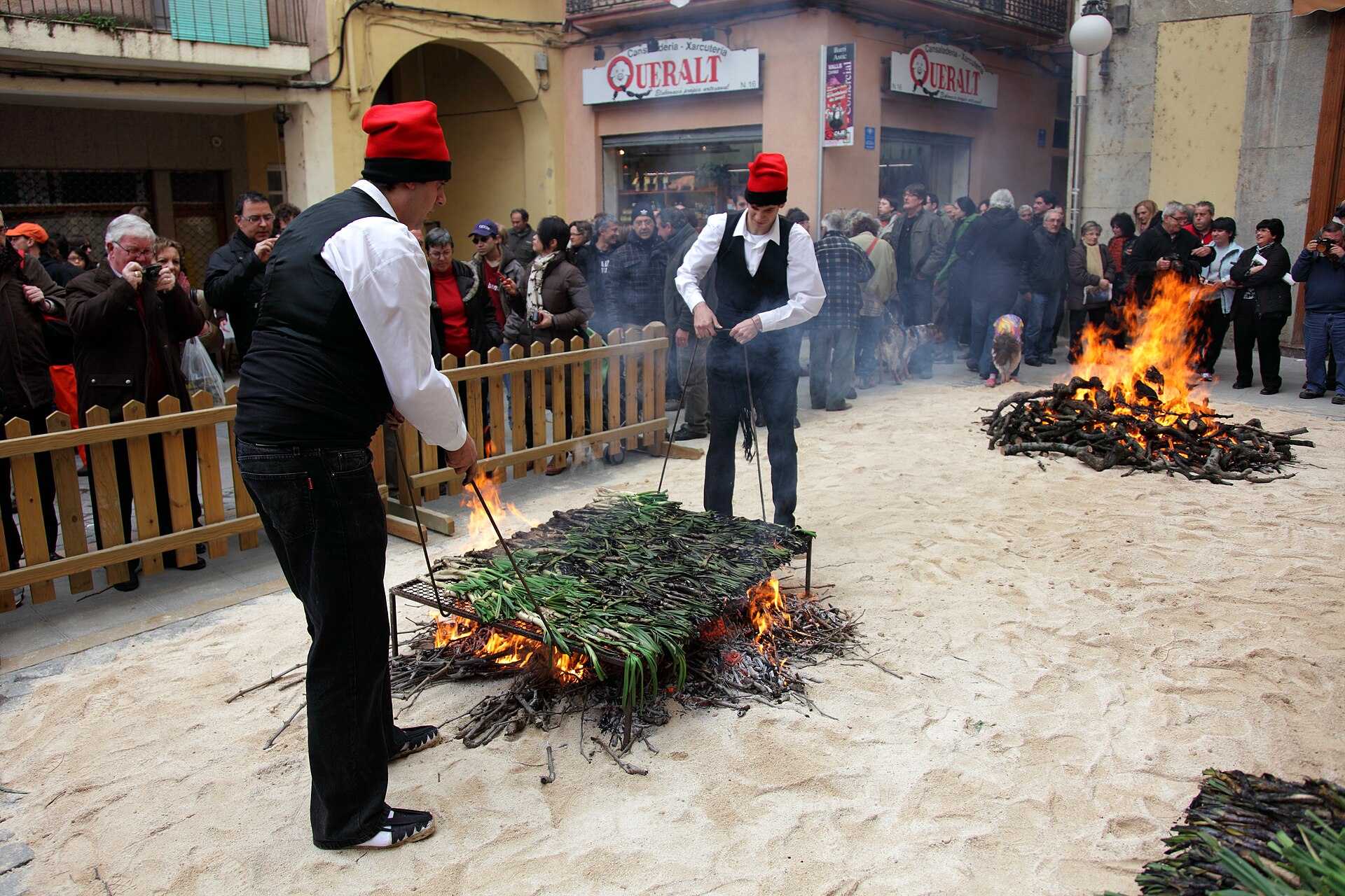 Two men in red Catalan barretina caps grilling bundles of calcots over an open fire of vine cuttings in a street in Valls