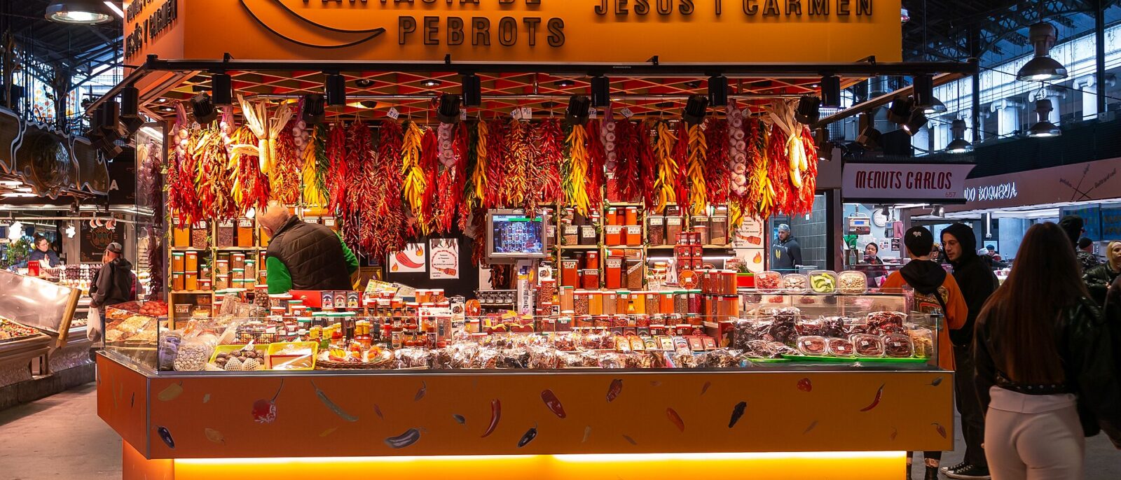 A brightly lit stall in La Boqueria market hung with strings of red and yellow chillies, garlic and jars of spices