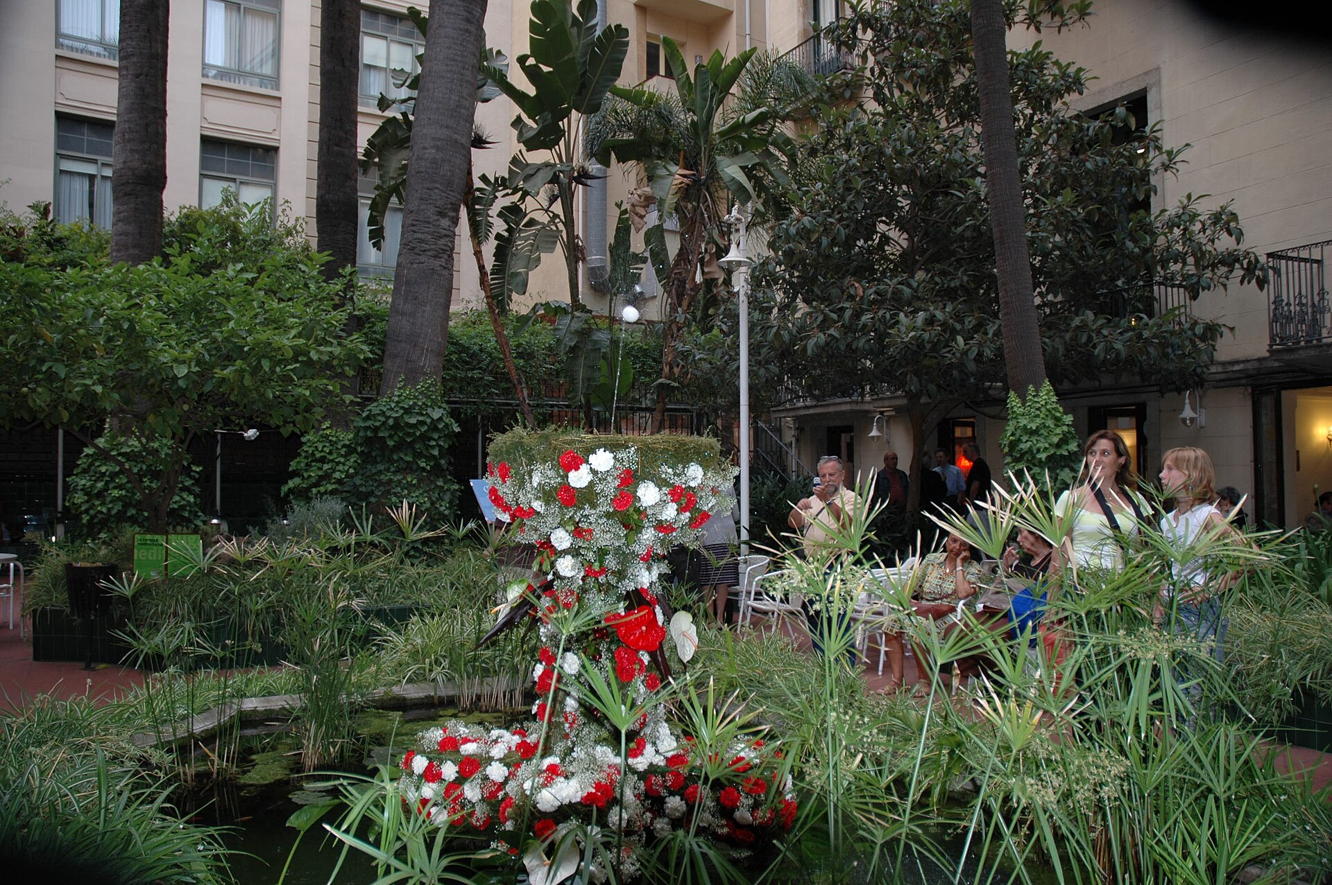 A flower-covered fountain in the garden courtyard of the Ateneu Barcelones with visitors watching l'Ou com balla