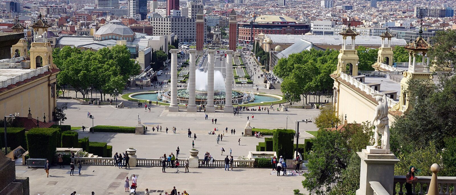 The Magic Fountain of Montjuic running in daylight, seen from the steps of the Palau Nacional, with the four columns, Placa d'Espanya and Tibidabo beyond
