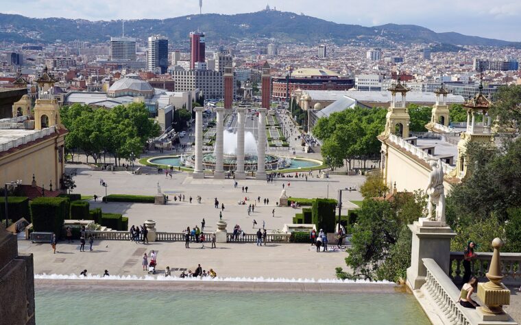 The Magic Fountain of Montjuic running in daylight, seen from the steps of the Palau Nacional, with the four columns, Placa d'Espanya and Tibidabo beyond