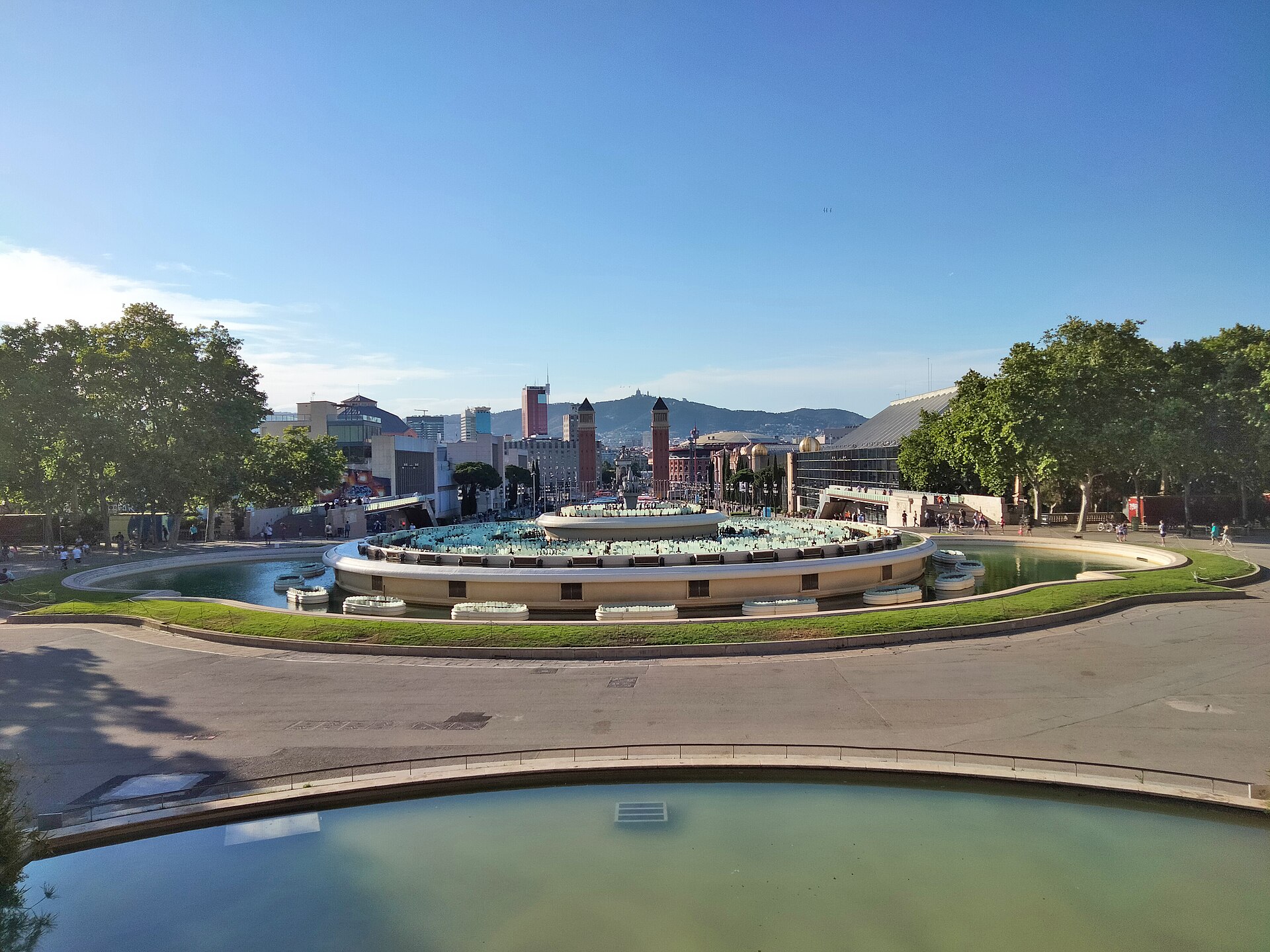 The Magic Fountain of Montjuic switched off in daylight, with its empty basin and the Venetian towers of Placa d'Espanya beyond