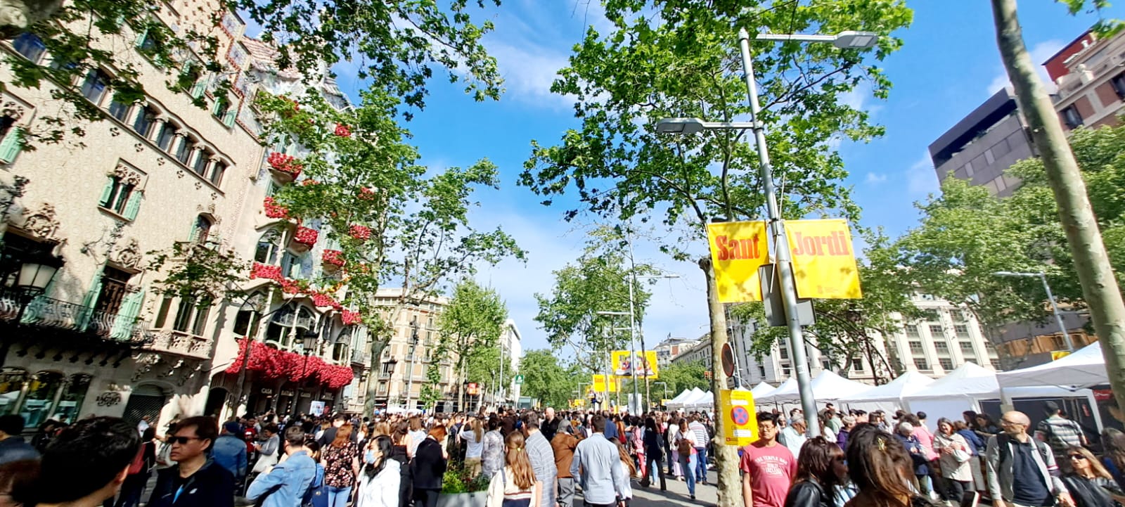 Crowds walking between book and rose stalls on a traffic-free Passeig de Gracia on Sant Jordi, with Casa Batllo's balconies covered in red roses