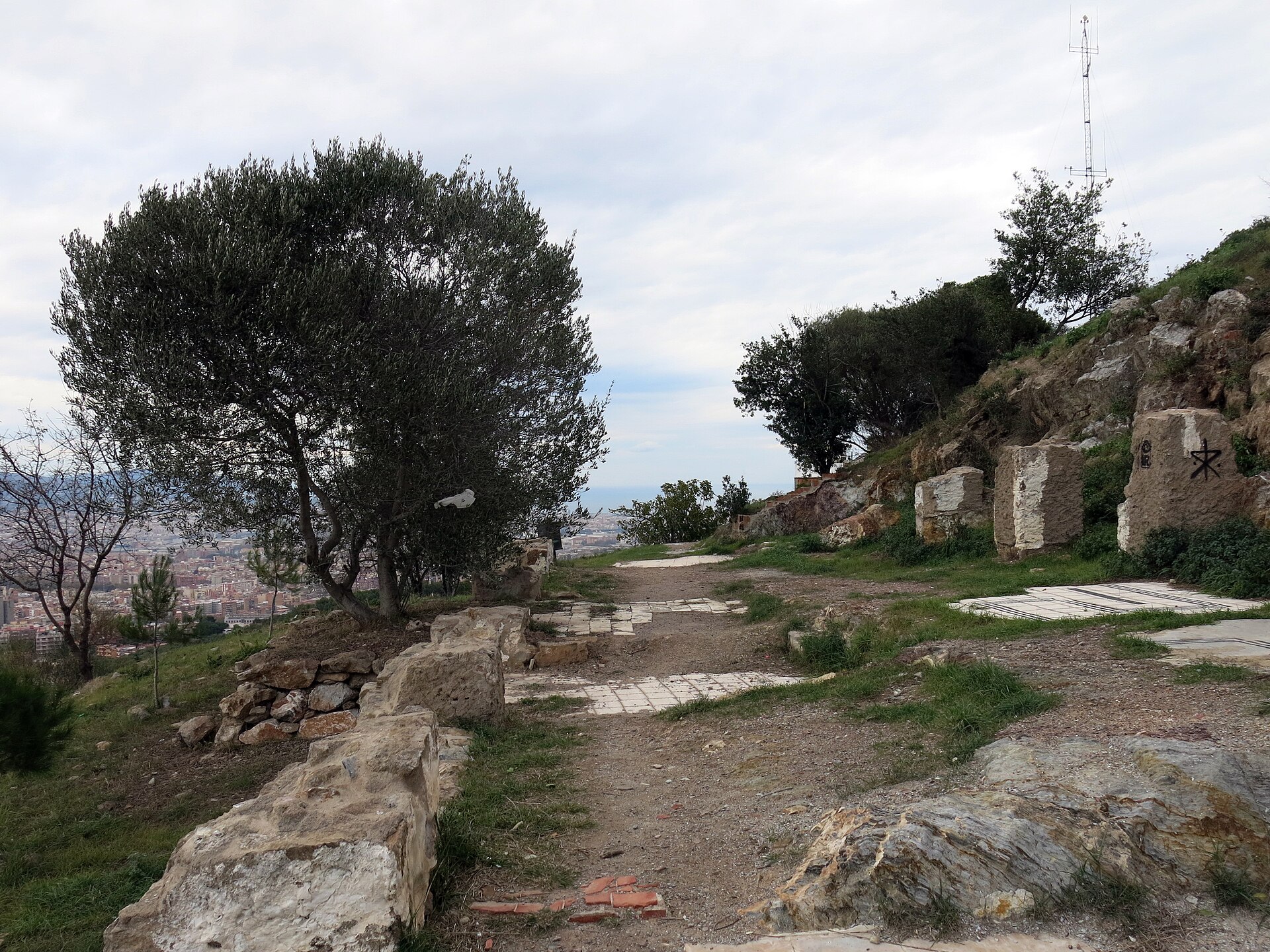 A dirt path on the summit of the Turo de la Rovira lined with the tiled floors of demolished shacks and stone blocks, with an olive tree and the city below