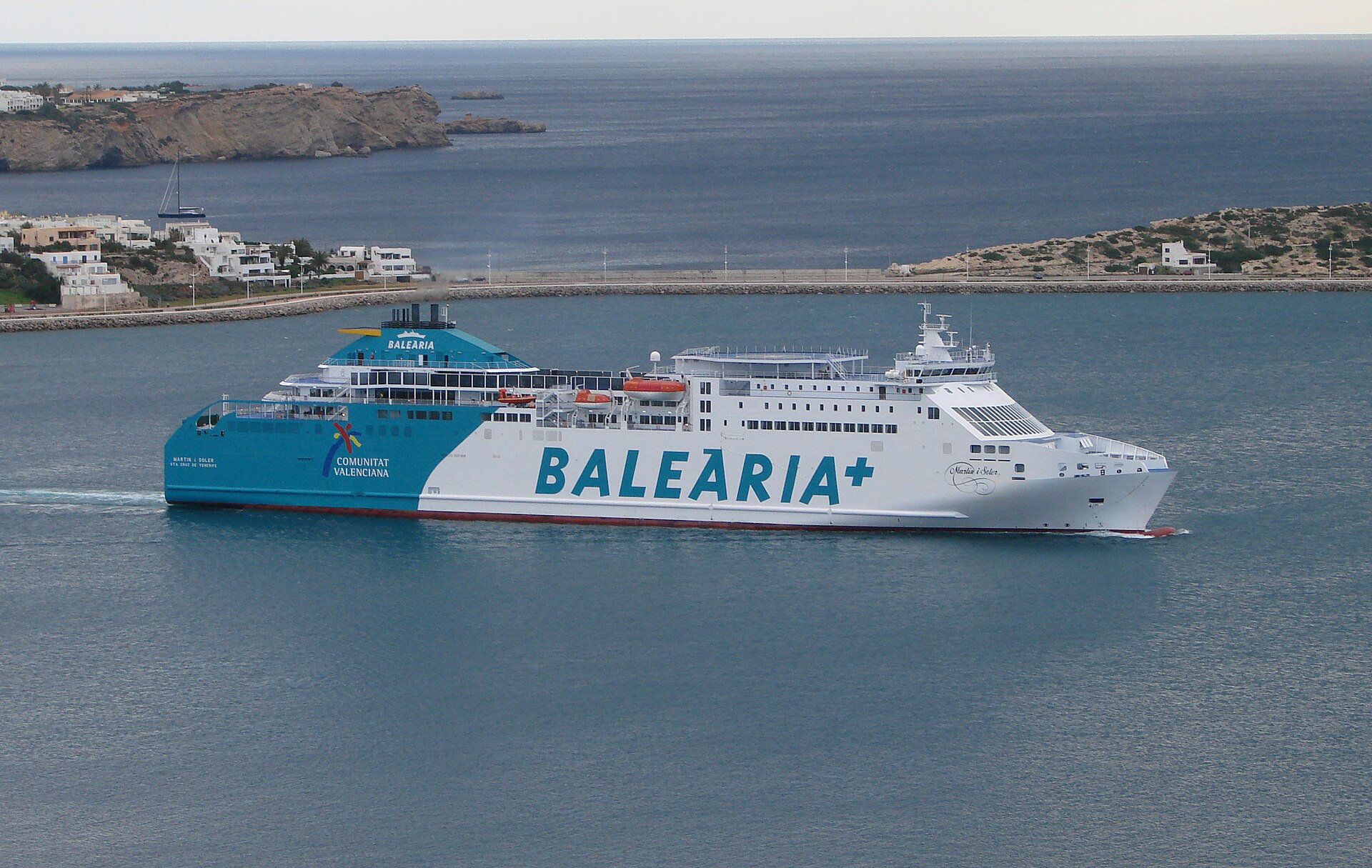 A large white and turquoise Balearia ferry sailing into Ibiza harbour, with the coast and houses behind