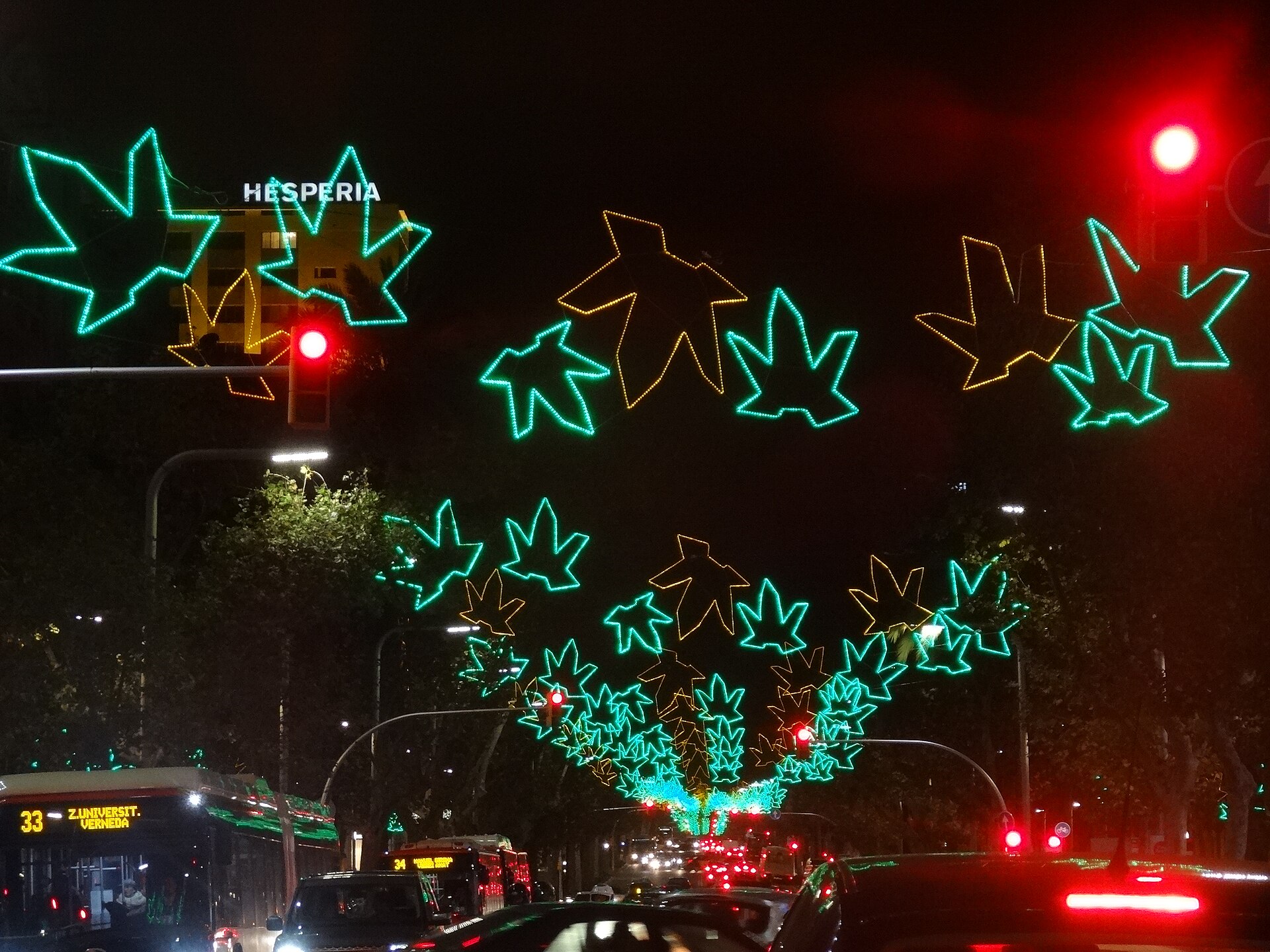 Christmas lights shaped like green and gold leaves strung above a wide Barcelona avenue at night, with buses and traffic below