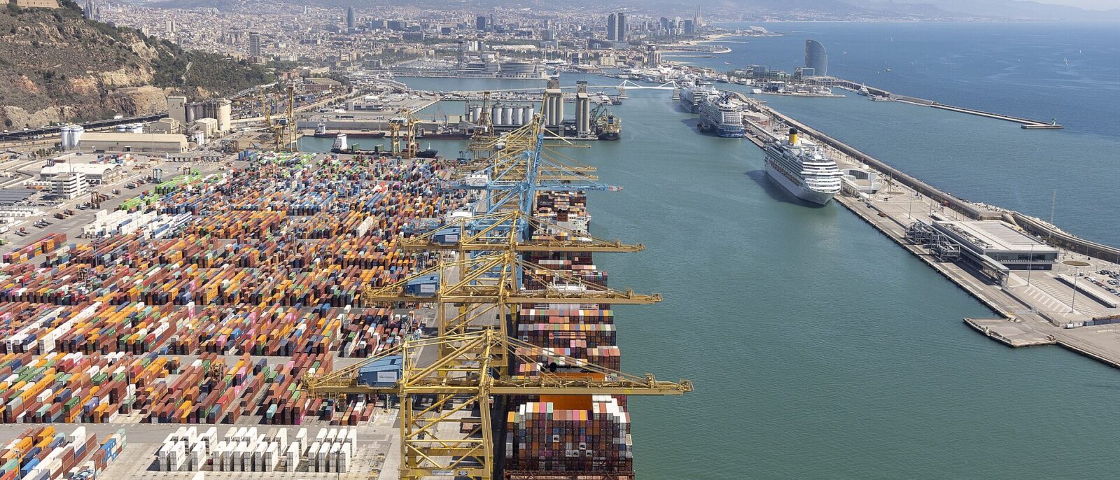 Aerial view of Barcelona's port, with the container terminal on the left and cruise ships berthed along the Moll Adossat on the right, the city and Montjuic behind