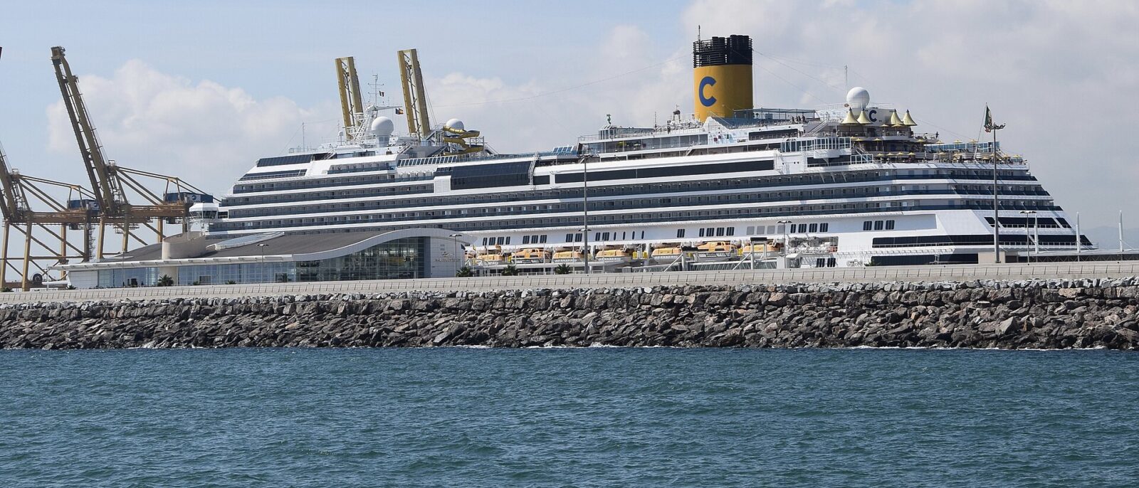 A Costa cruise ship berthed beside a terminal building on the Moll Adossat in Barcelona, with container cranes on the left