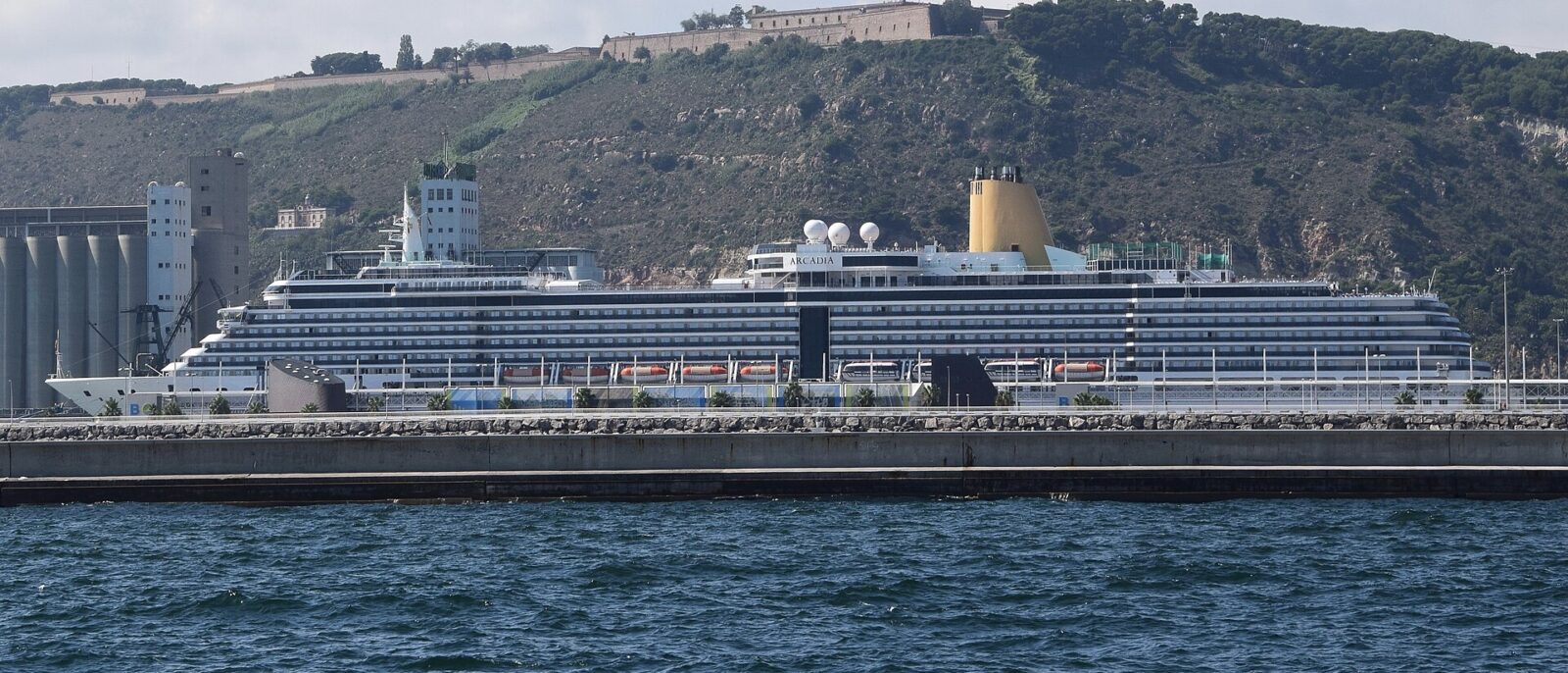 A large cruise ship berthed at the Moll Adossat in Barcelona, with grain silos to the left and Montjuic Castle on the hill above