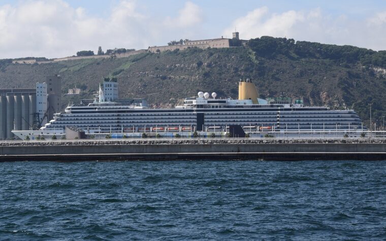 A large cruise ship berthed at the Moll Adossat in Barcelona, with grain silos to the left and Montjuic Castle on the hill above