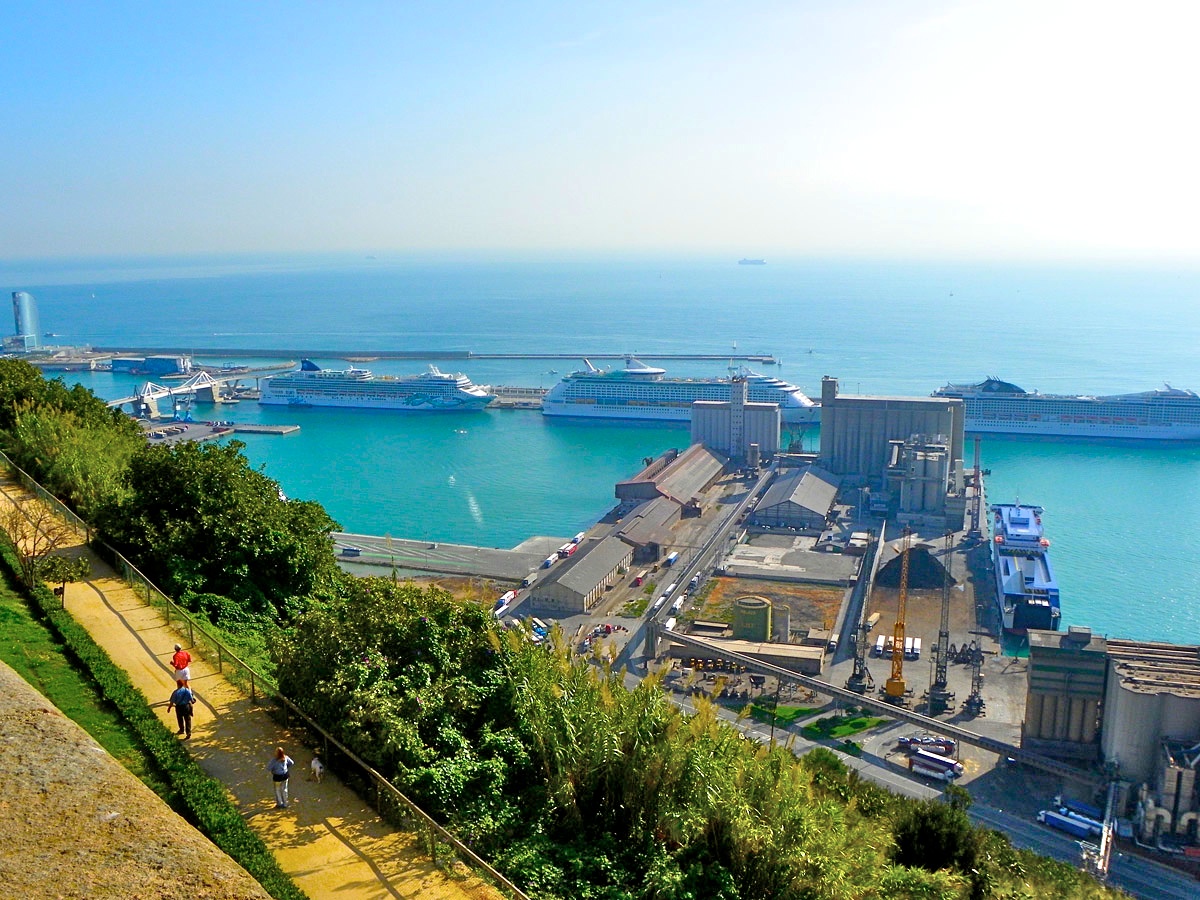 Three large cruise ships berthed at the Moll Adossat in Barcelona, seen from the gardens of Montjuic, with grain silos and port buildings in the foreground