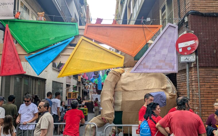 Giant coloured triangles and a hessian figure hung over Carrer de Jesus in Gracia, with visitors at the barriers below, during the 2026 Festa Major de Gracia