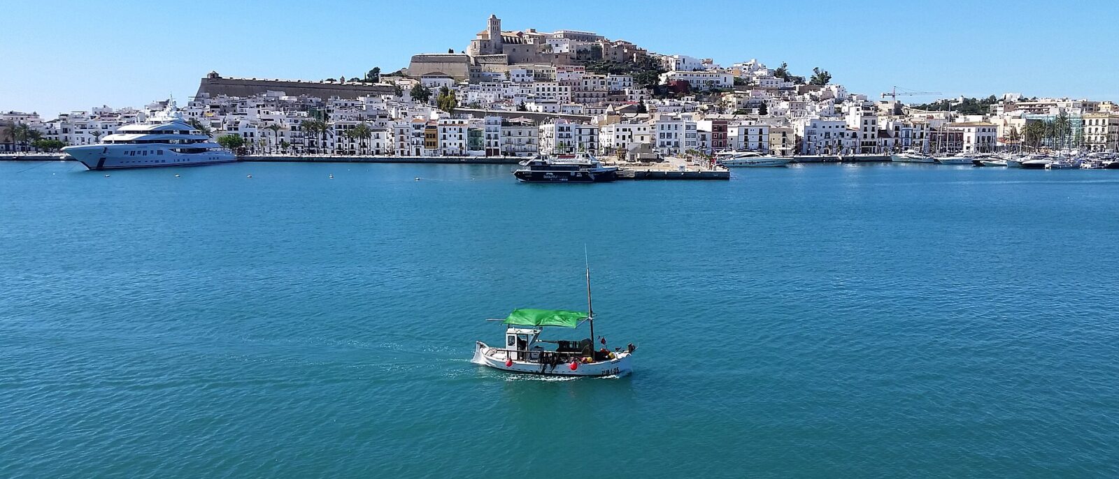 The white houses and walls of Dalt Vila rising above Ibiza harbour, with a small fishing boat crossing the bay in the foreground