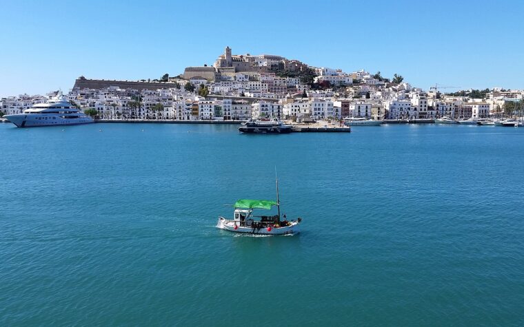 The white houses and walls of Dalt Vila rising above Ibiza harbour, with a small fishing boat crossing the bay in the foreground