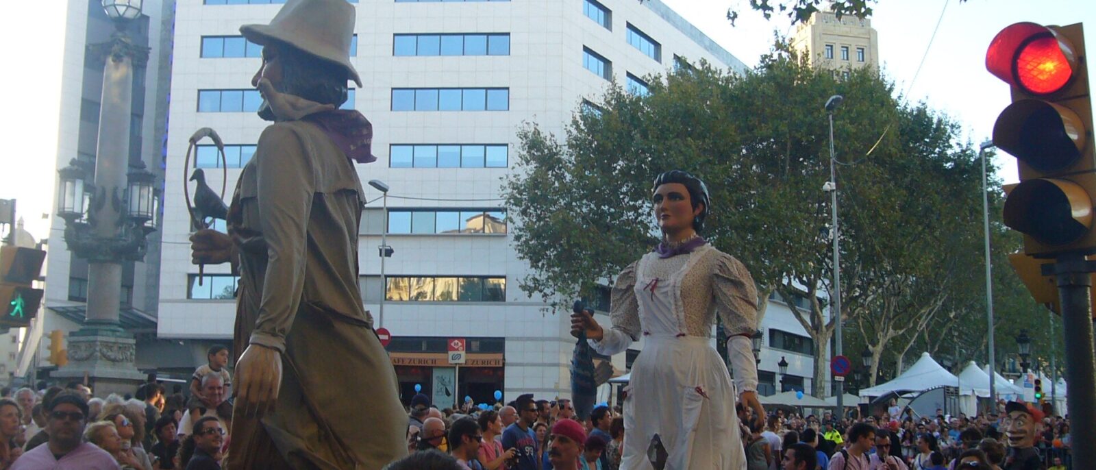 Two festival giants, a bearded man in a hat and a woman in a white dress, parading above a crowd in the Placa de Catalunya during La Merce