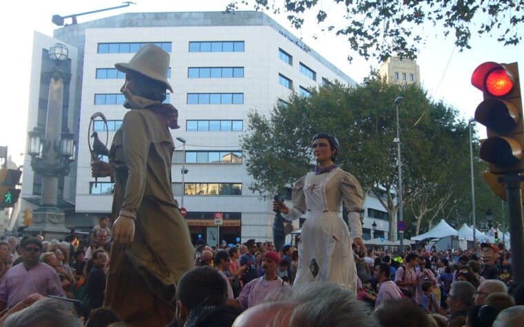 Two festival giants, a bearded man in a hat and a woman in a white dress, parading above a crowd in the Placa de Catalunya during La Merce