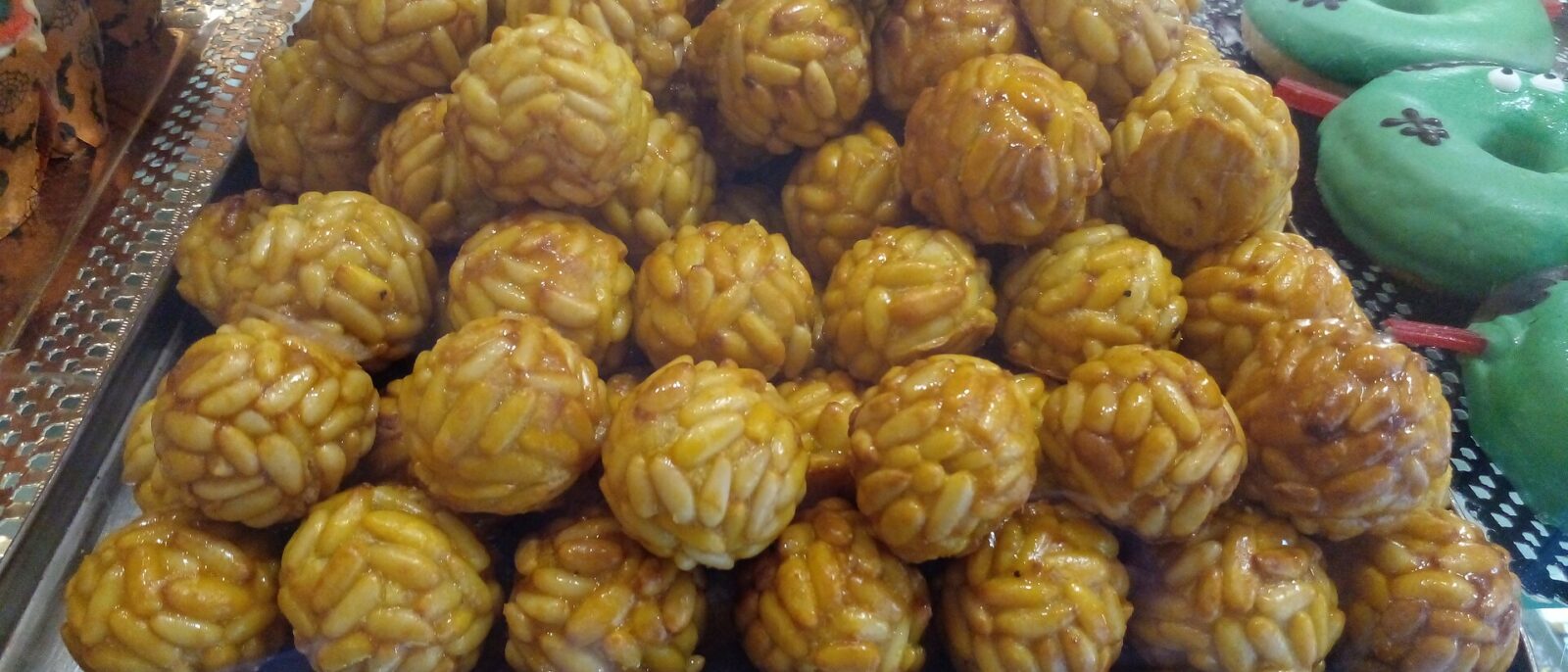 A tray of pine-nut panellets in a pastry shop window, with Halloween-decorated doughnuts beside them