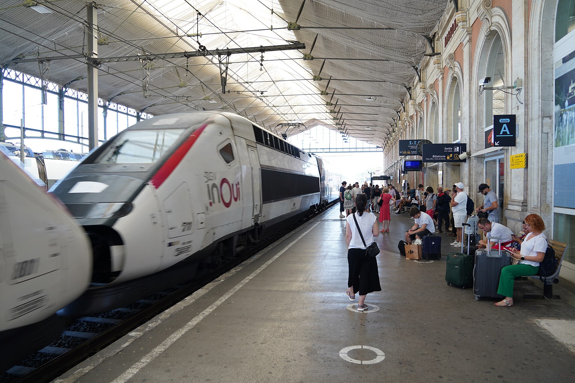 A TGV inOui high-speed train arriving at Narbonne station, with passengers and luggage waiting on the platform under the old station roof