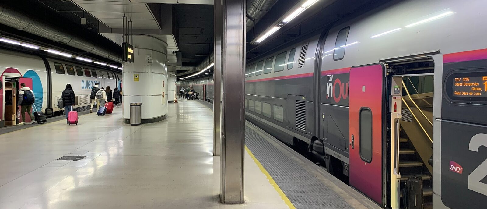 A double-deck TGV inOui train at a platform in Barcelona Sants, its door screen showing Girona and Paris Gare de Lyon, with a Ouigo train on the next track