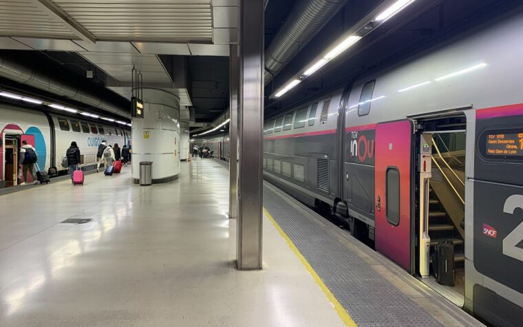 A double-deck TGV inOui train at a platform in Barcelona Sants, its door screen showing Girona and Paris Gare de Lyon, with a Ouigo train on the next track